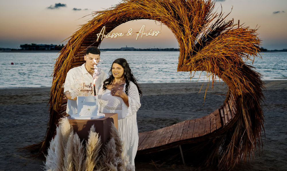 Couple celebrates engagement at beach with heart-shaped decor and cake during sunset.