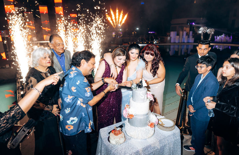 Group celebrating a birthday with a cake and fireworks in the background at a nighttime event by the pool.