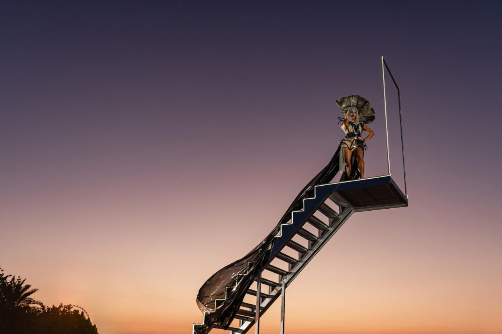 Person in a dramatic costume stands on a raised platform at sunset, showcasing avant-garde fashion against a twilight sky.