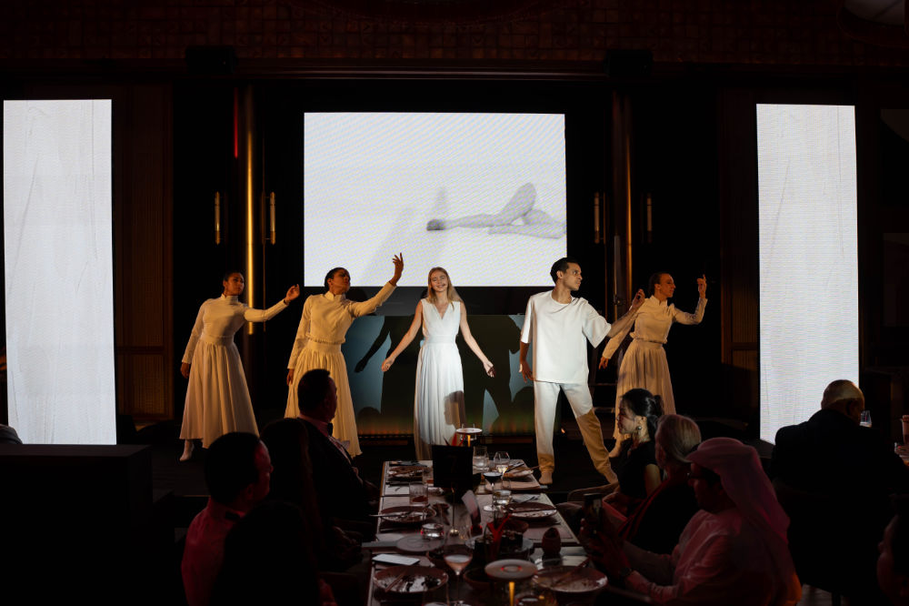 Performers in white outfits on stage during a vibrant live show with audience seating in the foreground.
