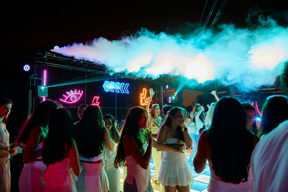 Group of people enjoying a vibrant neon party under colorful lights and smoke effects at night.