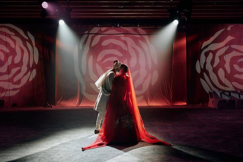 Couple dances under spotlight, surrounded by abstract floral backdrops, with bride in a stunning red gown.