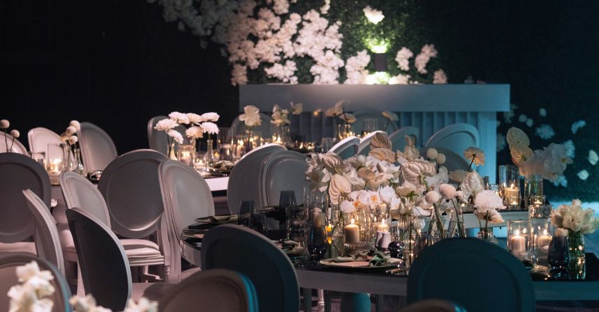 Elegant event setup with white flowers and candles on tables, surrounded by chic chairs and floral wall decor.