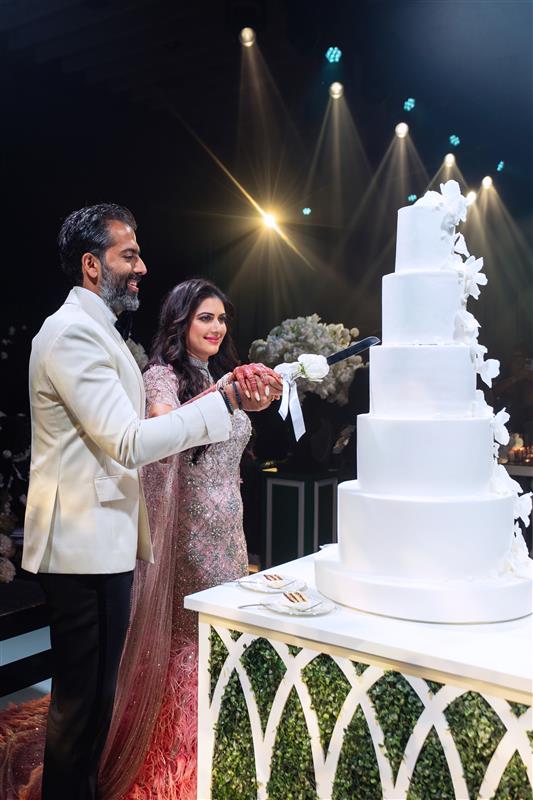 A couple in formal attire cuts a tall, white wedding cake adorned with flowers under spotlights.