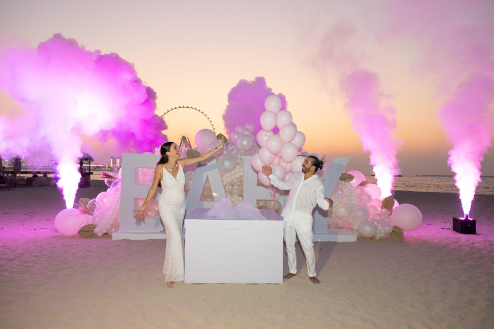 Couple celebrates gender reveal on beach at sunset, surrounded by pink smoke and balloons spelling BABY.