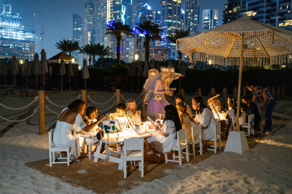 Children painting at a decorated beachside table under a parasol at night, with city skyline in the background.